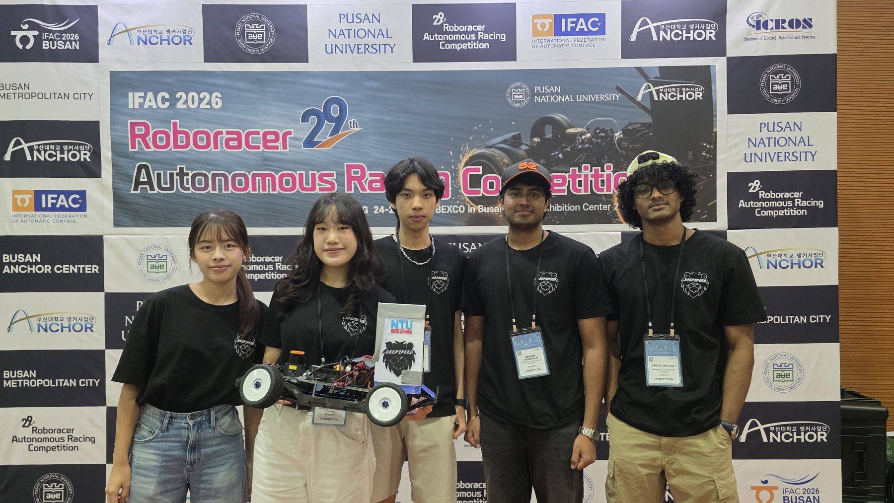 Five NTU DeepSpeed members with their car in front of the IFAC 2026 RoboRacer backdrop